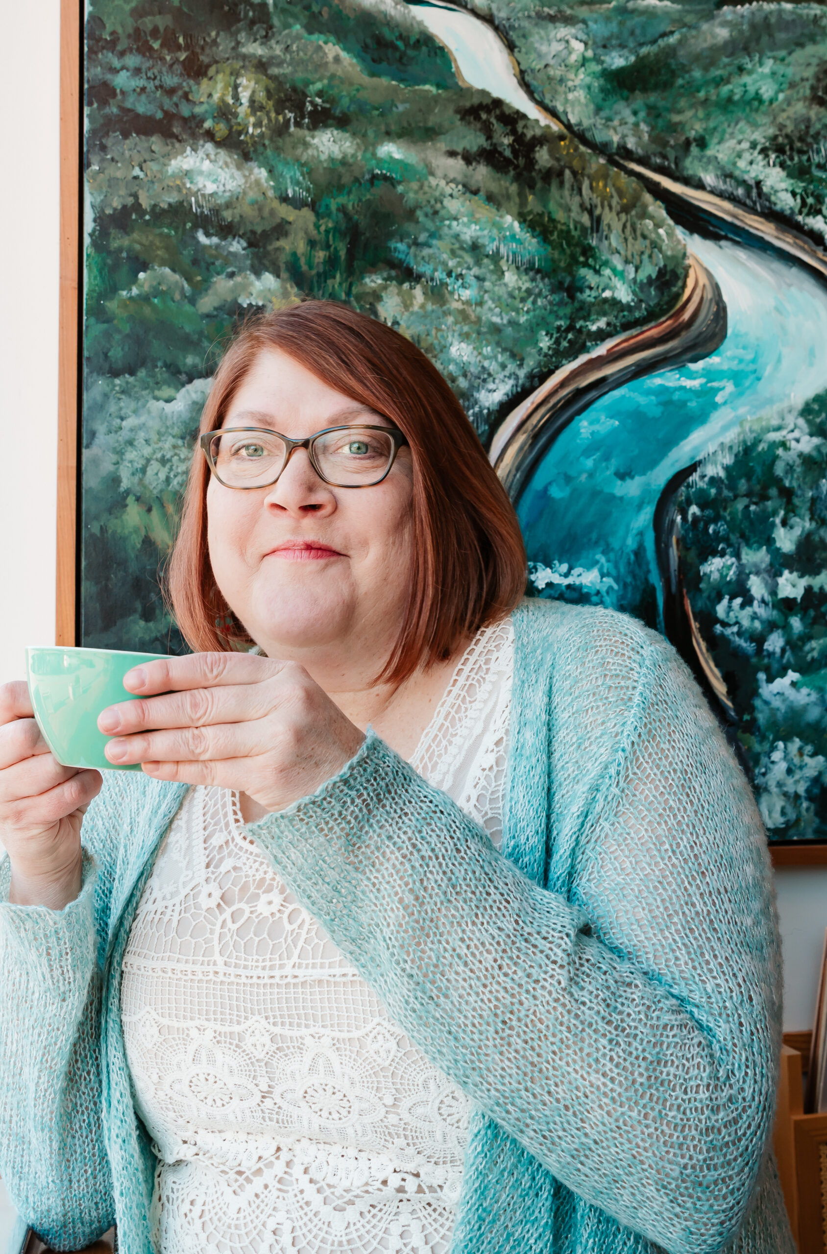 woman sitting in front of a painting of a river and forest and dringing coffee